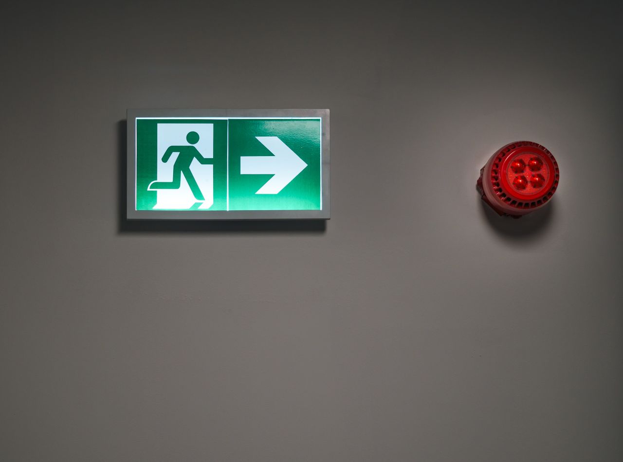 Home A brightly lit exit sign and alarm in an indoor corridor, indicating emergency escape route.